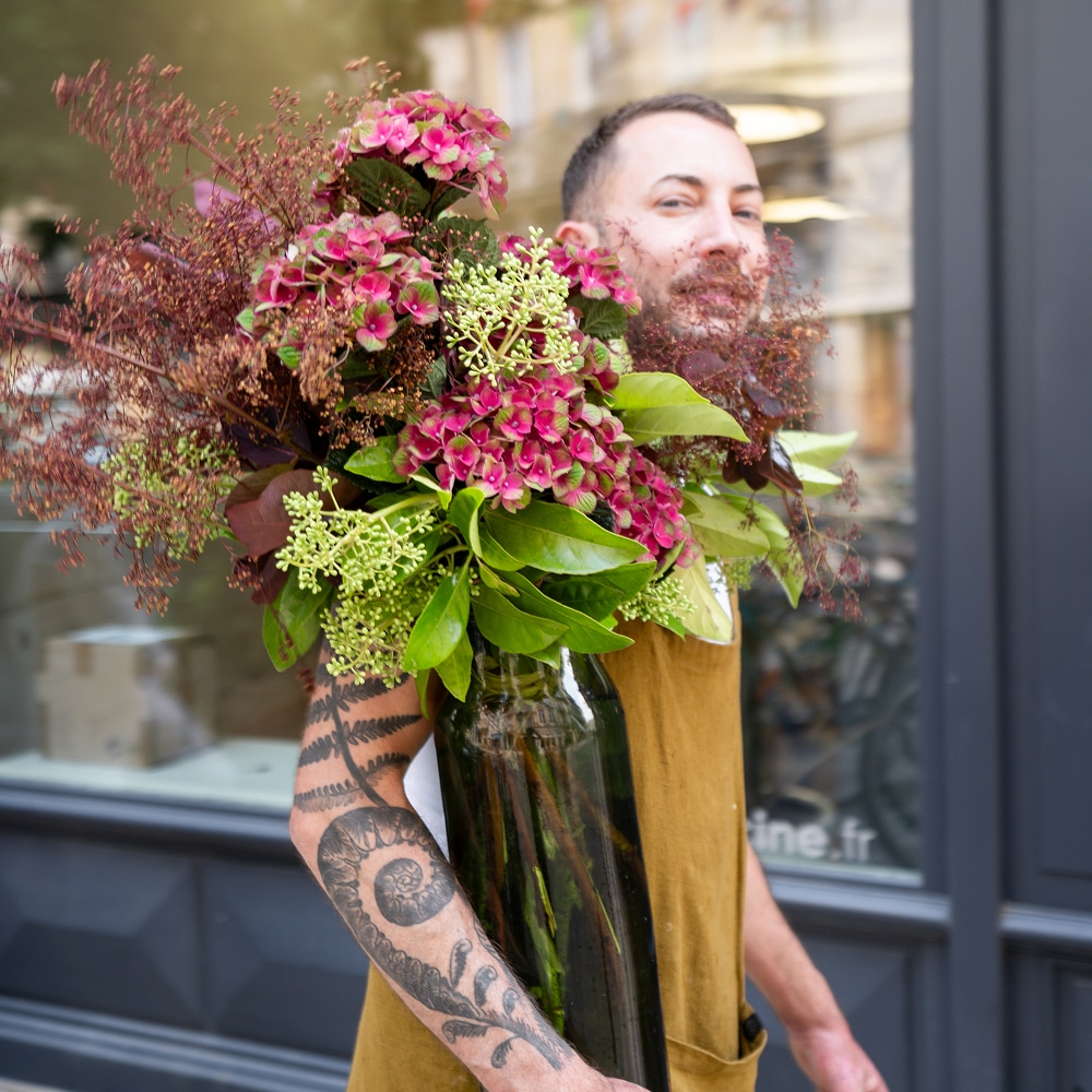 Fleuriste souriant, bras tatoué, en tablier marron, porte un grand bouquet d'hortensias roses et baies vertes. Fenêtre "...tine.fr".