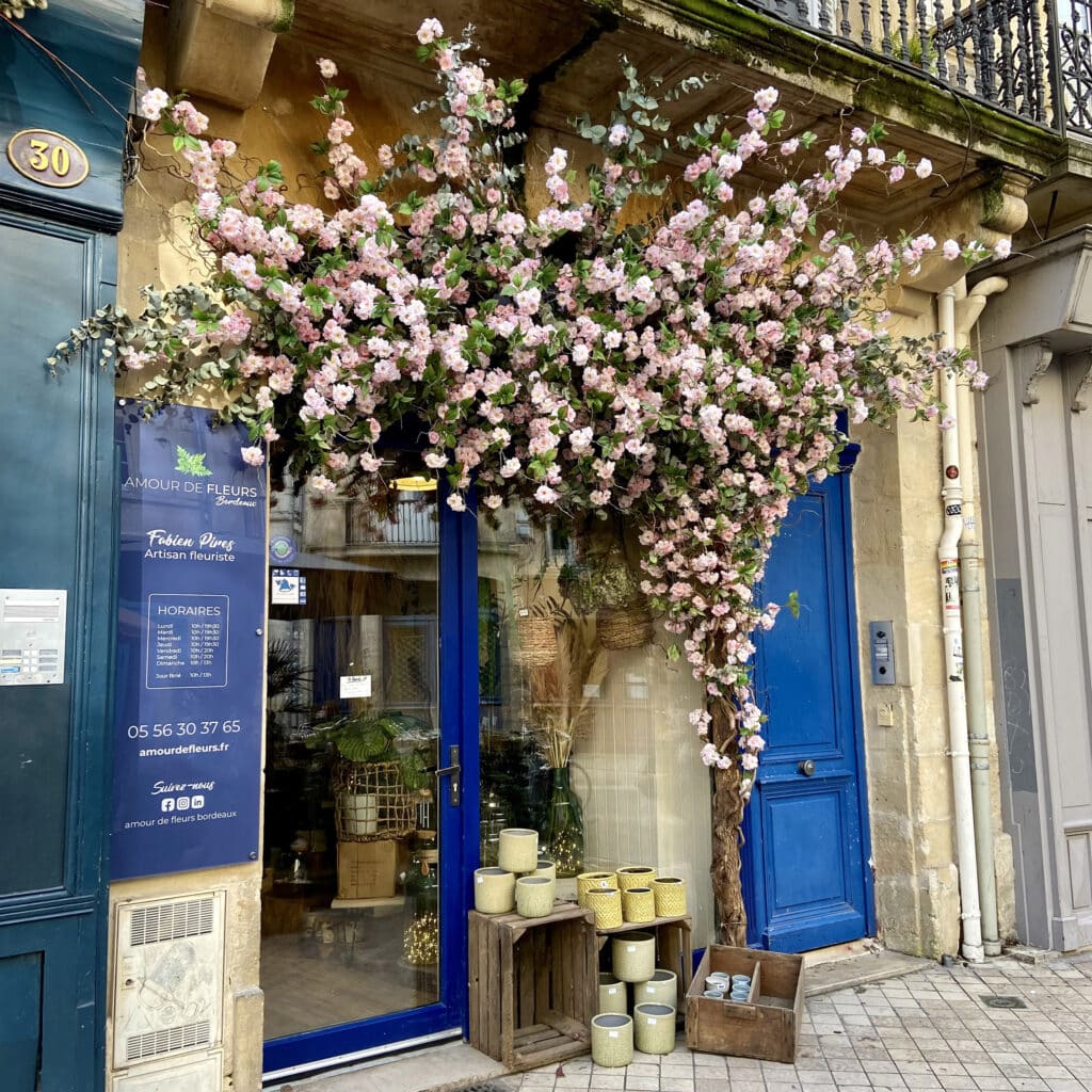 Façade du fleuriste Amour de Fleurs Bordeaux, 30. Entrée bleue ornée de fleurs roses et eucalyptus. Détails Fabien Pires, horaires et contact affichés.