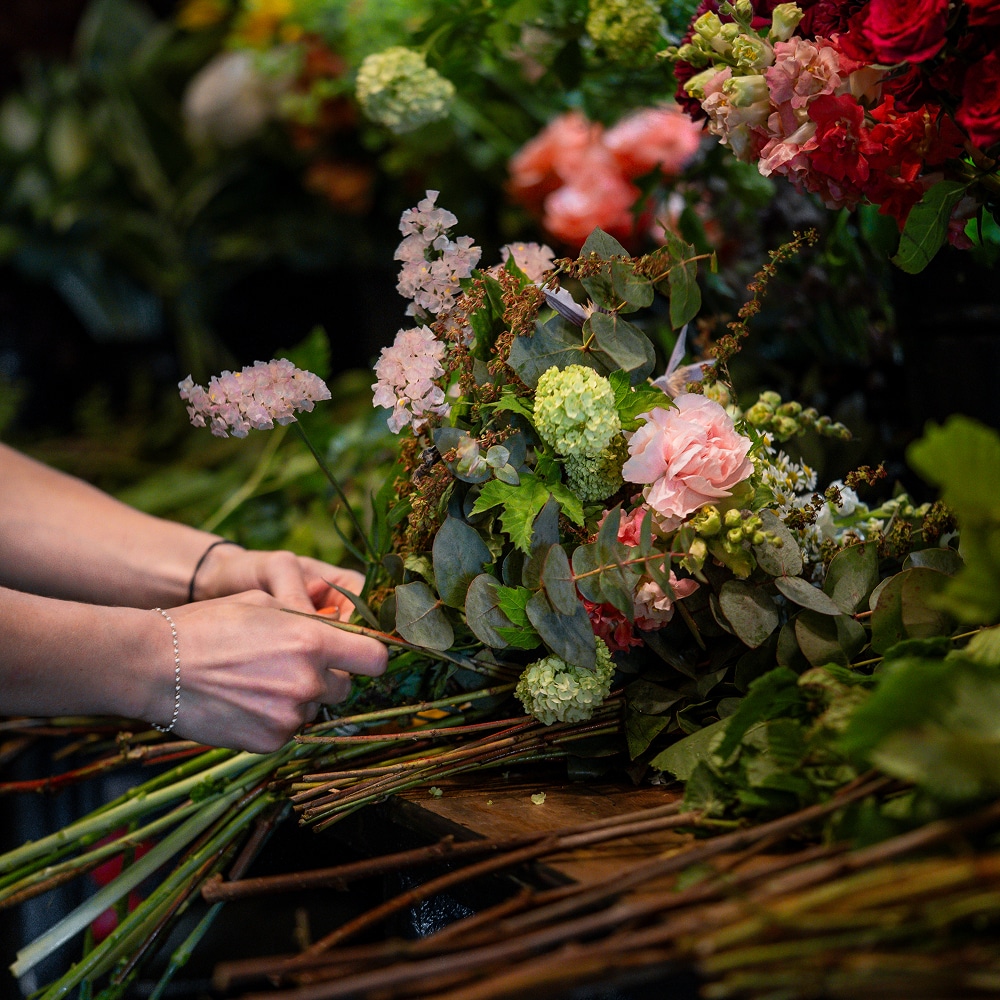 Mains d'un floriste composant un bouquet frais : œillets roses, petites fleurs pâles, eucalyptus et hydrangéas verts, sur un établi en bois.
